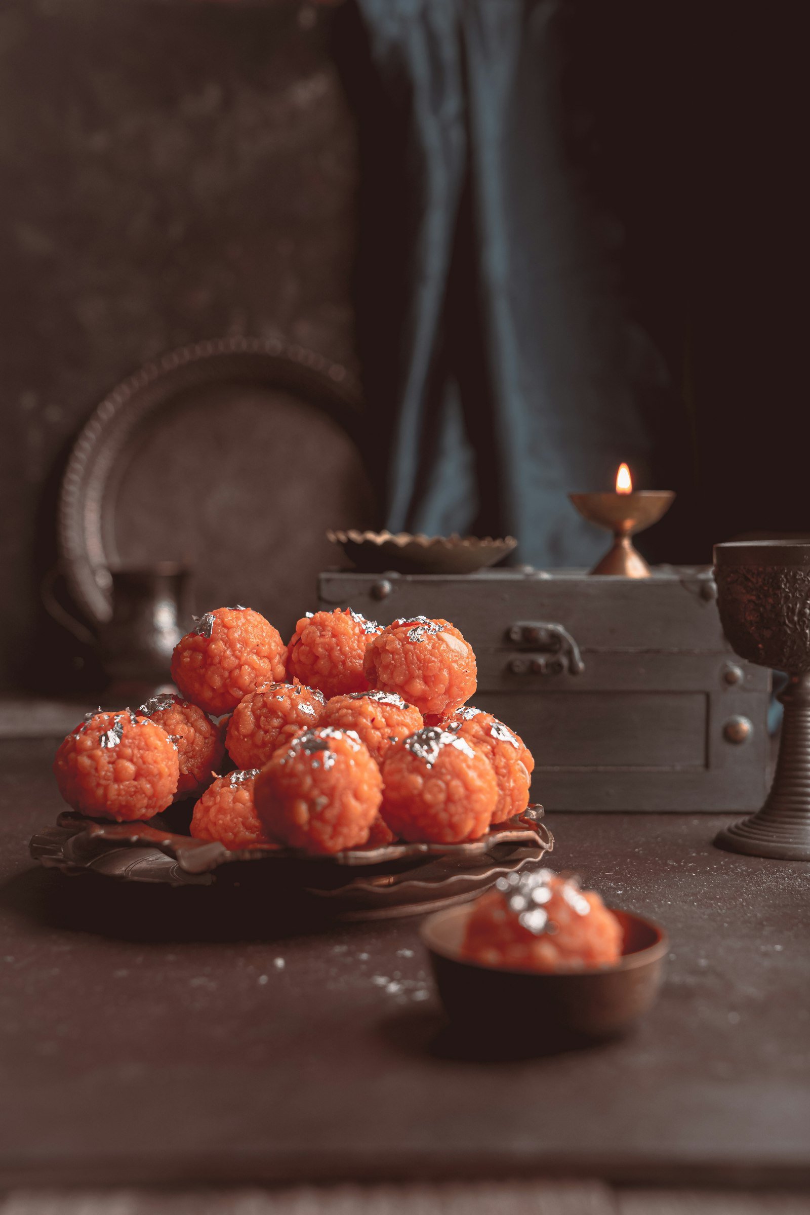Orange laddoos on a dark table with a lit brass diya behind them