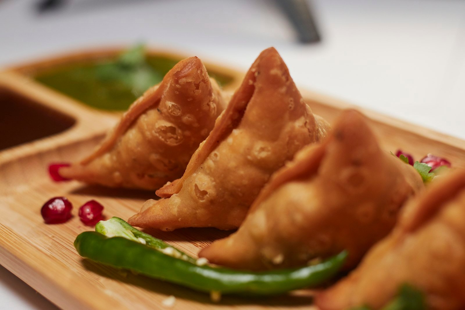 Golden fried samosas on a wooden board with green chillies and pomegranate